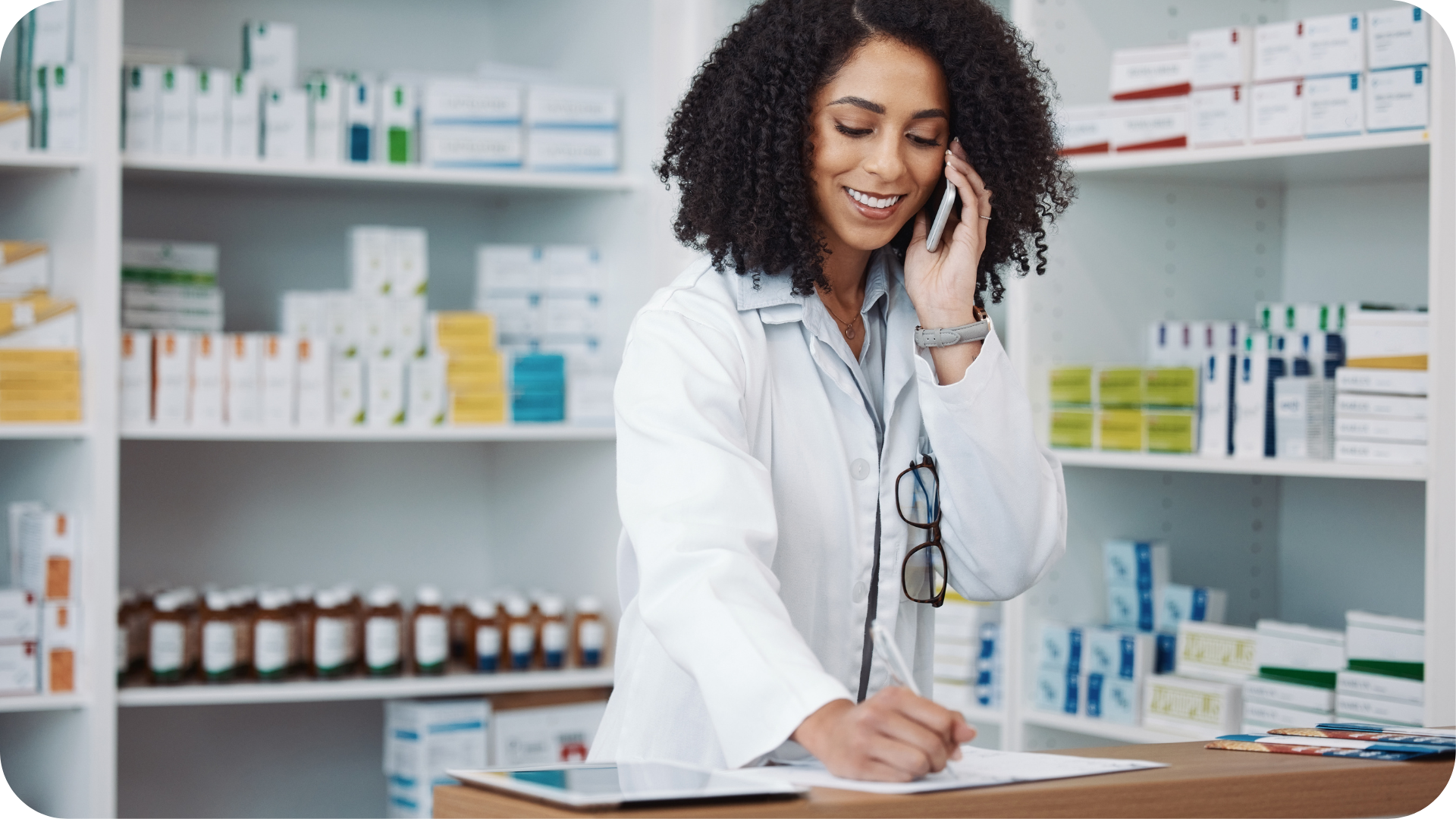 Female pharmacist on the phone taking notes with medications on shelves behind her.