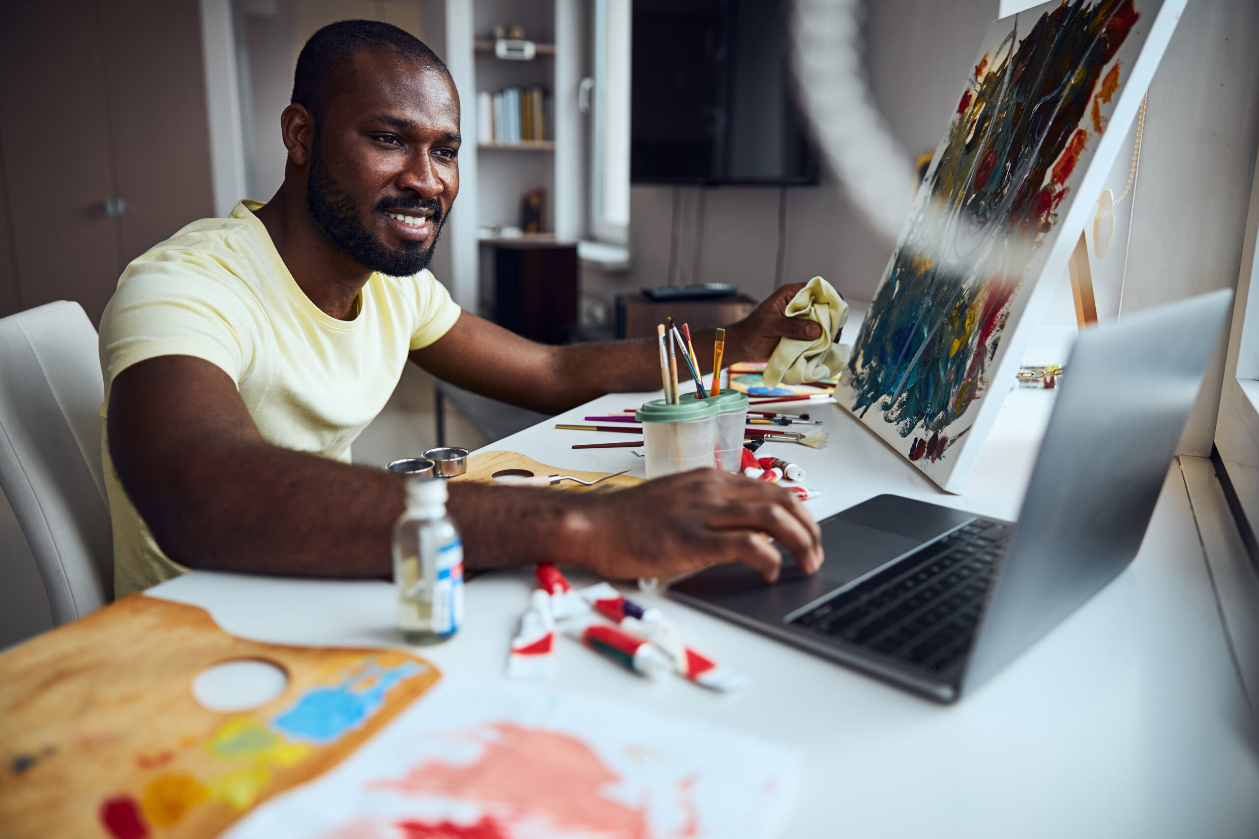 Mixed-race creator moving his hand on a touchpad while watching the laptop display and sitting opposite to a picture